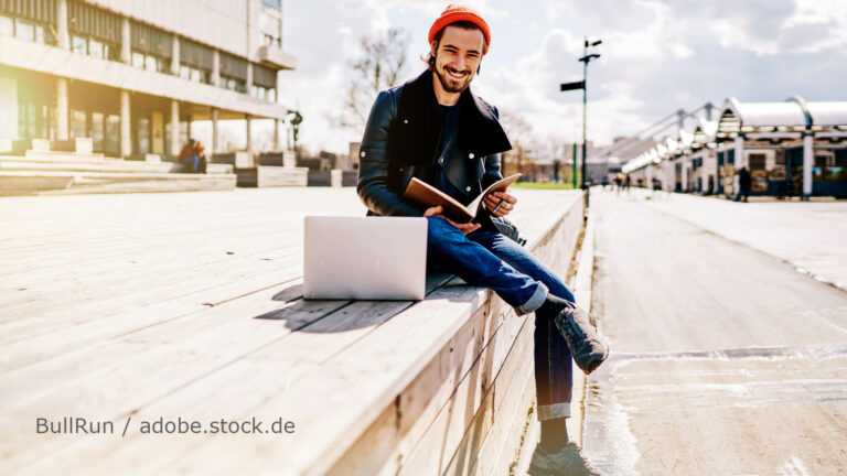 Mann mit roter Mütze sitzt mit Laptop und Buch auf einer Mauer