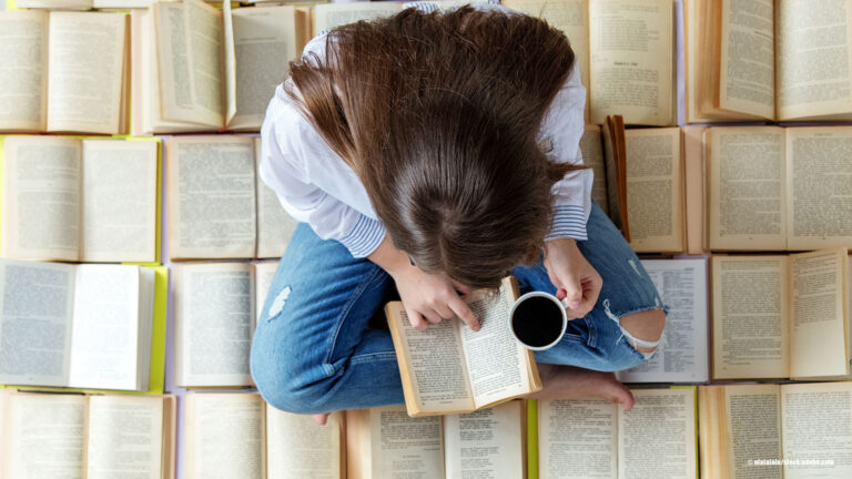 Frau sitzt auf aufgeklappten Büchern und schaut mit einer Kaffeetasse in der Hand in ein Buch auf dem Schoß.