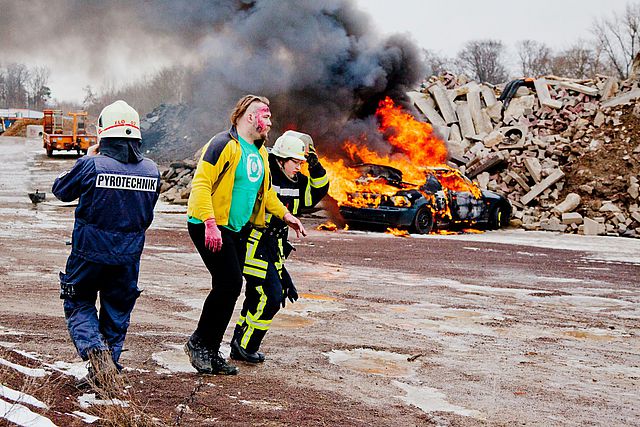 Ein Feuerwehrmann führt eine verletzte Person von der Unfallstelle fort. Im Hintergrund brennt etwas.|Ein Feuerwehrmann führt eine verletzte Person von der Unfallstelle fort. Im Hintergrund brennt etwas.|Zwei Frauen führen erste Hilfe bei einem Menschen durch